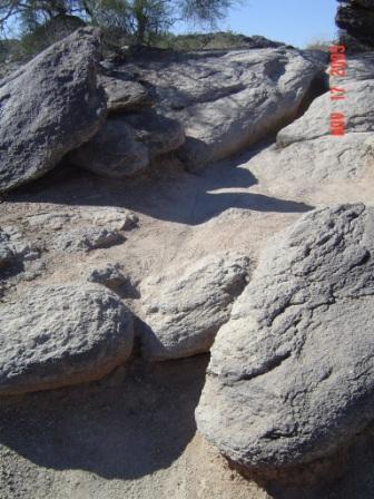 A rocky terrain with large boulders and sandy ground, under clear blue sky. The scene depicts natural stone formations and the surrounding landscape. South Mountain Park / National Trail mountain bike trail.