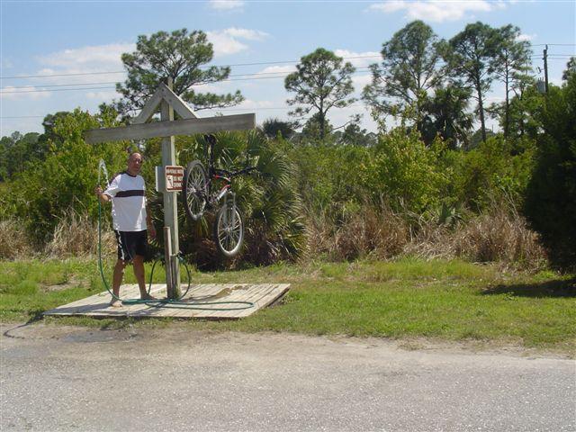 A man stands next to a bicycle repair station with a bike suspended above a wooden platform. The station is surrounded by lush greenery and tall trees under a bright blue sky. The sign on the station indicates that bicycle repair services are available. Caloosahatchee Regional Park mountain bike trail.