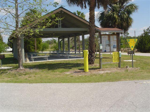 A covered picnic area with several picnic tables, surrounded by green grass and trees. There is a signboard with informational postings nearby, as well as a yellow post. The scene is bright and sunny, typical of an outdoor recreational space. Caloosahatchee Regional Park mountain bike trail.