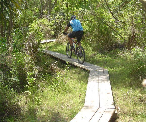 A person riding a mountain bike on a wooden pathway through a lush, green forested area, surrounded by dense vegetation and trees. Caloosahatchee Regional Park mountain bike trail.