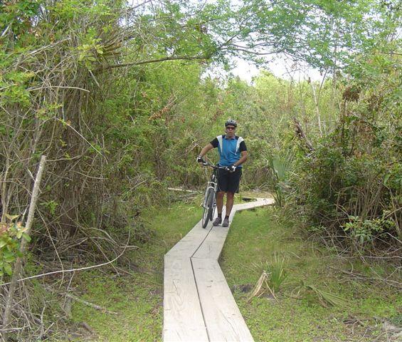 A person in a cycling outfit stands next to a mountain bike on a wooden path surrounded by lush greenery and dense vegetation. The path winds through a natural environment, leading into the woods. Caloosahatchee Regional Park mountain bike trail.