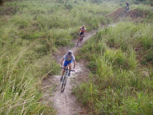 Three mountain bikers riding on a dirt trail through a grassy landscape. The foreground features two riders, one in blue and the other in orange, navigating a curve, while a third rider can be seen in the background on a different path. The area is surrounded by tall grasses and a natural, earthy setting. Caloosahatchee Regional Park mountain bike trail.