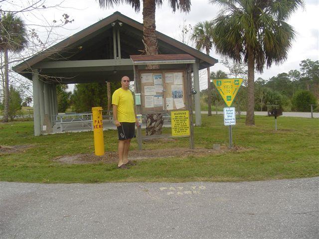 A man in a yellow shirt stands near an informational kiosk at a park, with a covered pavilion in the background. The kiosk displays various notices and warnings, while nearby signs indicate park rules and directions. Palm trees are visible in the surrounding greenery. Caloosahatchee Regional Park mountain bike trail.