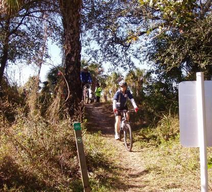 A person riding a mountain bike on a dirt trail surrounded by trees and greenery. In the background, additional cyclists can be seen ascending a slope. A signpost is visible in the foreground, indicating the trail. The scene is bright and sunny, typical of an outdoor biking environment. Caloosahatchee Regional Park mountain bike trail.
