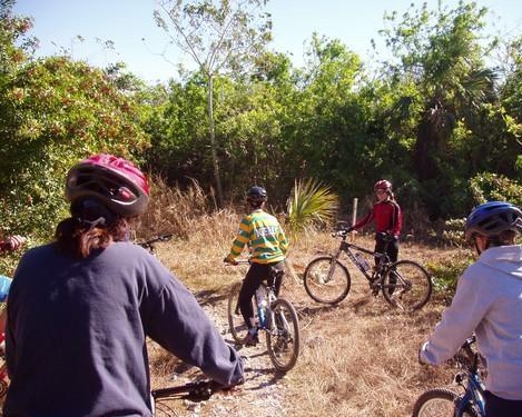 A group of mountain bikers wearing helmets is gathered on a dirt path surrounded by greenery. The participants are engaged with one another, with one person in a striped jersey facing the group while others are on their bikes, ready to ride. The scene depicts a sunny day outdoors in a natural setting. Caloosahatchee Regional Park mountain bike trail.