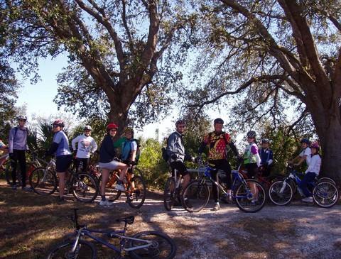 A group of cyclists posing for a photo on a dirt path surrounded by trees. They are dressed in various cycling gear and helmets, with several bicycles parked nearby. The scene captures a sunny day in a natural outdoor setting. Caloosahatchee Regional Park mountain bike trail.