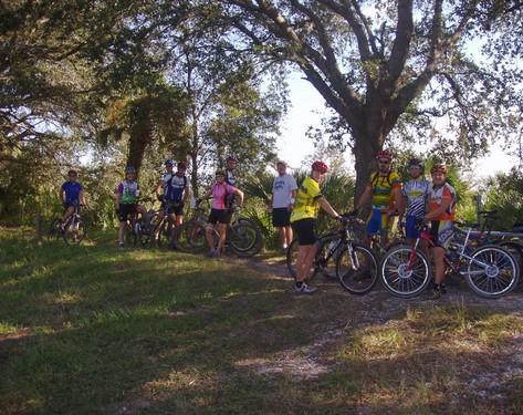 A group of ten cyclists, wearing colorful jerseys and helmets, stands together near their bicycles in a lush green outdoor setting. The scene features a mix of male and female riders, with trees and foliage in the background, suggesting a recreational biking event in nature. Caloosahatchee Regional Park mountain bike trail.