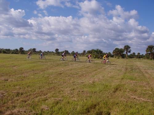 A group of six cyclists riding in single file through a grassy field under a partly cloudy sky. Caloosahatchee Regional Park mountain bike trail.
