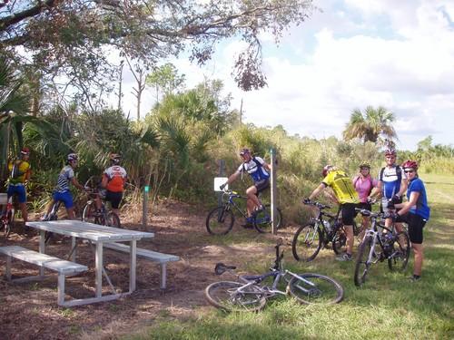 A group of eight cyclists gathered at a picnic area in a natural setting, featuring a table and bicycles scattered around. The cyclists are wearing colorful jerseys and helmets, surrounded by lush greenery and palm trees under a partly cloudy sky. Caloosahatchee Regional Park mountain bike trail.