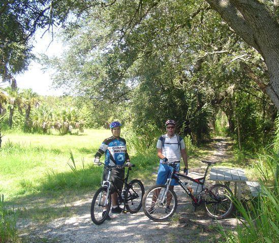 Two mountain bikers standing next to their bikes on a dirt path surrounded by greenery and trees. One biker is wearing a blue and black cycling jersey and a helmet, while the other is in a white shirt and jeans. A picnic table is visible nearby in the natural setting. Caloosahatchee Regional Park mountain bike trail.
