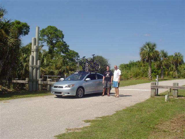 Two men stand next to a silver car parked on a sunny road surrounded by palm trees and lush greenery. A bicycle is mounted on the roof of the car. In the background, there is a wooden structure and a clear blue sky. The scene suggests a recreational outing. Caloosahatchee Regional Park mountain bike trail.