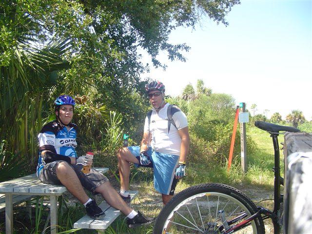 Two cyclists resting at a picnic table in a lush outdoor setting. One person is sitting on the table with a drink, wearing a blue cycling jersey and shorts, while the other stands nearby, dressed in a white shirt and cycling gear. A bicycle is positioned in the foreground, and trees and greenery are visible in the background. Caloosahatchee Regional Park mountain bike trail.