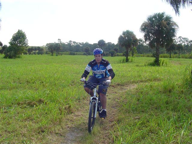 A person in cycling gear rides a mountain bike along a dirt path through a grassy field, surrounded by trees and palm plants under a bright sky. Caloosahatchee Regional Park mountain bike trail.