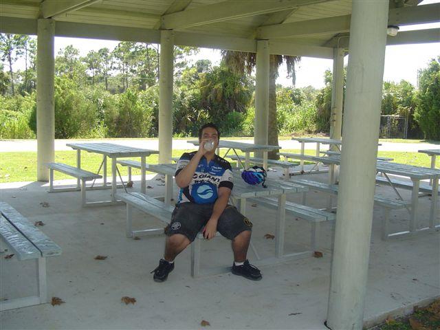 A person sitting on a picnic bench under a pavilion, wearing a blue and white cycling jersey and black shorts, while drinking from a bottle. The surrounding area features greenery and palm trees in the background. Caloosahatchee Regional Park mountain bike trail.