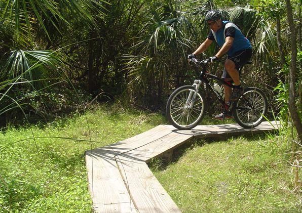 A person riding a mountain bike on a wooden trail through a lush, green forest. The rider is focused and wearing a helmet, navigating a zigzag section of the pathway surrounded by tall grasses and tropical plants. Caloosahatchee Regional Park mountain bike trail.
