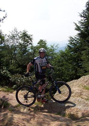 A mountain biker in a cycling jersey stands next to a bicycle on rocky terrain, surrounded by greenery and trees under a bright sky. Kitsuma mountain bike trail.