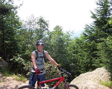 A man in a tank top and helmet stands beside a mountain bike on a rocky trail, surrounded by trees and greenery. The sun is shining in a clear blue sky, indicating a bright, outdoor setting. Kitsuma mountain bike trail.