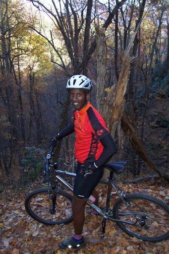 A person wearing a red and black cycling jersey and helmet stands next to a mountain bike on a trail surrounded by autumn foliage. Trees with colorful leaves are visible in the background, indicating a mountainous or wooded area. Kitsuma mountain bike trail.