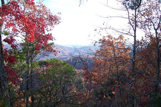 A scenic view of a mountainous landscape during autumn, featuring trees with vibrant red and orange leaves, visible layers of hills in the background, and a clear sky. Kitsuma mountain bike trail.