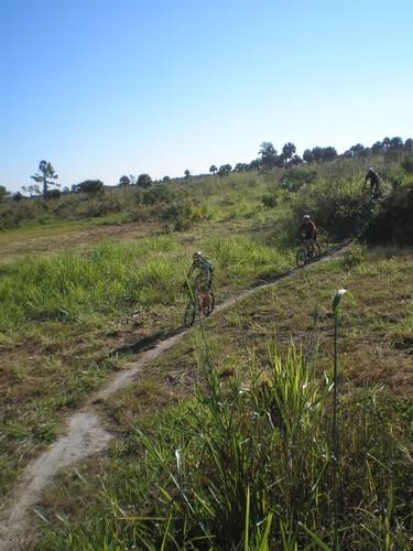 Bicyclists riding along a dirt path through a grassy field under a clear blue sky. Caloosahatchee Regional Park mountain bike trail.