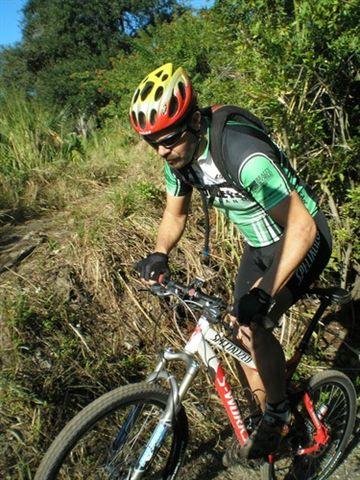 A cyclist navigating a dirt trail lined with greenery, wearing a green and black cycling jersey and a brightly colored helmet. The cyclist is focused and in motion on a mountain bike. Caloosahatchee Regional Park mountain bike trail.
