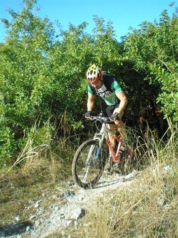 Alt text: A mountain biker wearing a helmet and cycling gear navigates a rocky trail surrounded by lush greenery and tall grass on a sunny day. Caloosahatchee Regional Park mountain bike trail.