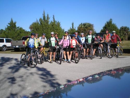 A group of cyclists wearing helmets and colorful jerseys poses for a photo with their mountain bikes in a parking area surrounded by trees on a clear day.  Caloosahatchee Regional Park mountain bike trail.