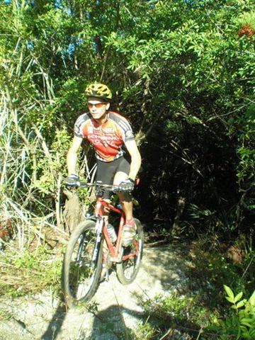 A cyclist in a yellow helmet rides a red mountain bike along a narrow trail surrounded by dense greenery. The scene captures the rider navigating through a natural, wooded area, showcasing the adventure of mountain biking. Caloosahatchee Regional Park mountain bike trail.