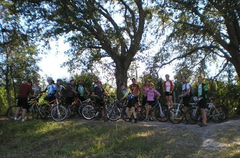 A group of cyclists posing with their bicycles in a natural outdoor setting, surrounded by trees and greenery. Caloosahatchee Regional Park mountain bike trail.