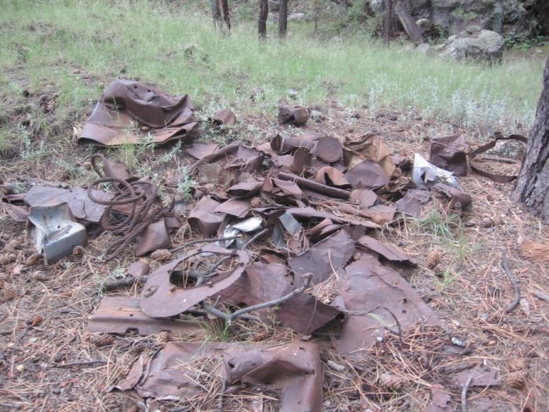 A pile of rusted metal debris and scrap materials scattered on the ground in a forested area, surrounded by grass and pine needles. Schultz Creek mountain bike trail.