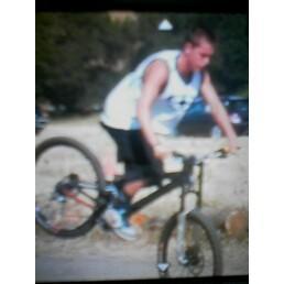 A person riding a mountain bike, performing a trick with the front wheel raised off the ground, on a dirt trail surrounded by greenery. The rider is wearing a sleeveless shirt and shorts. Caloosahatchee Regional Park mountain bike trail.