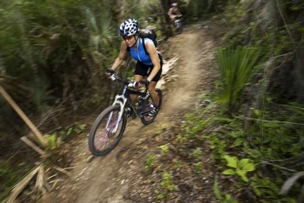 A person wearing a blue athletic tank top and a helmet rides a mountain bike on a dirt trail surrounded by lush greenery. The rider leans forward, demonstrating speed and agility, while another cyclist follows in the background. Caloosahatchee Regional Park mountain bike trail.