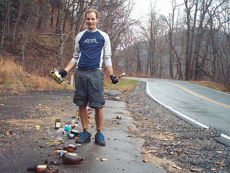 A man standing next to a rural road, holding a bottle and a piece of broken glass, with various discarded bottles scattered on the ground around him. Trees line the background on a cloudy day. Kitsuma mountain bike trail.