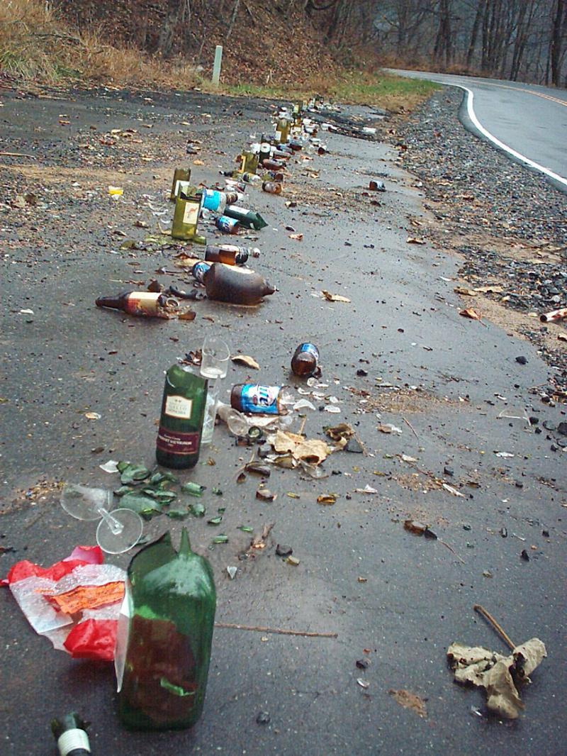 A long line of various glass and plastic bottles, including beer and liquor bottles, scattered along a paved road. The ground is littered with broken glass, leaves, and debris, indicating littering and environmental neglect. Trees and a curved road are visible in the background. Kitsuma mountain bike trail.