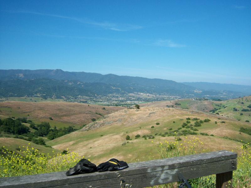 A scenic view of rolling hills and distant mountains under a clear blue sky, with a wooden bench in the foreground. Wildflowers and green vegetation are visible in the landscape, while a small town is nestled in the valley below. Santa Teresa Park mountain bike trail.
