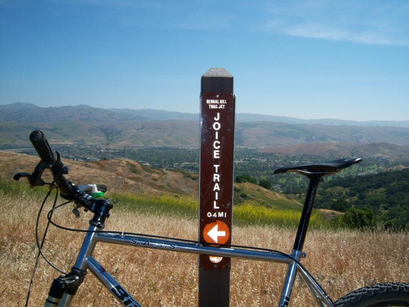 A mountain bike parked next to a trail sign indicating the Joice Trail, with a distance of 0.4 miles. The background features rolling hills and a clear blue sky, providing a scenic view of the landscape. Santa Teresa Park mountain bike trail.