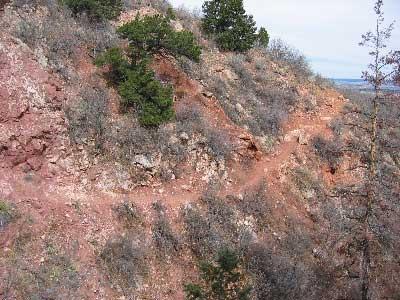 A narrow dirt trail winding along a rocky hillside covered with sparse vegetation and small green shrubs under a cloudy sky. Paul Intemann Memorial Trail mountain bike trail.