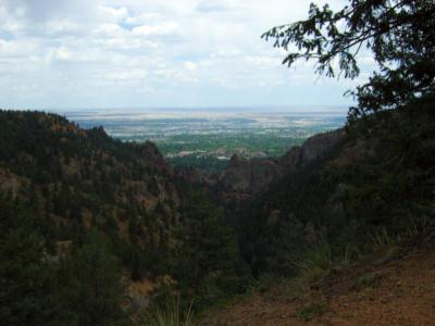A scenic view from a mountain overlook, showcasing a green valley and distant farmland, under a partly cloudy sky. Trees and rocky terrain frame the image, emphasizing the natural landscape. Columbine mountain bike trail.