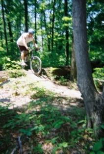 A person riding a mountain bike on a dirt trail in a lush green forest. The rider is in mid-air as they navigate a small jump, surrounded by trees and dense foliage. Kickapoo mountain bike trail.