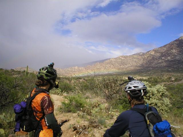 Two cyclists standing on a rocky trail, looking out over a scenic landscape featuring mountains and valleys under a partly cloudy sky. Vegetation, including cacti and shrubs, is visible in the foreground. 50-year Trail / Golder Ranch mountain bike trail.