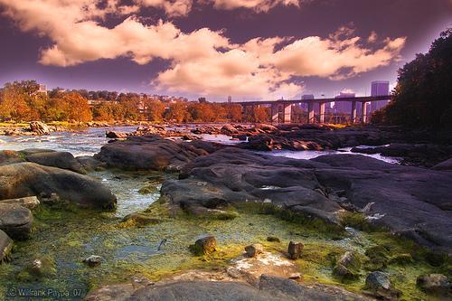 A scenic view of a river with rocky banks, featuring green and brown vegetation, under a dramatic sky filled with clouds. A bridge is seen in the background, with a city skyline partially visible. The vibrant colors of the landscape suggest a late afternoon or sunset setting. Belle Isle mountain bike trail.