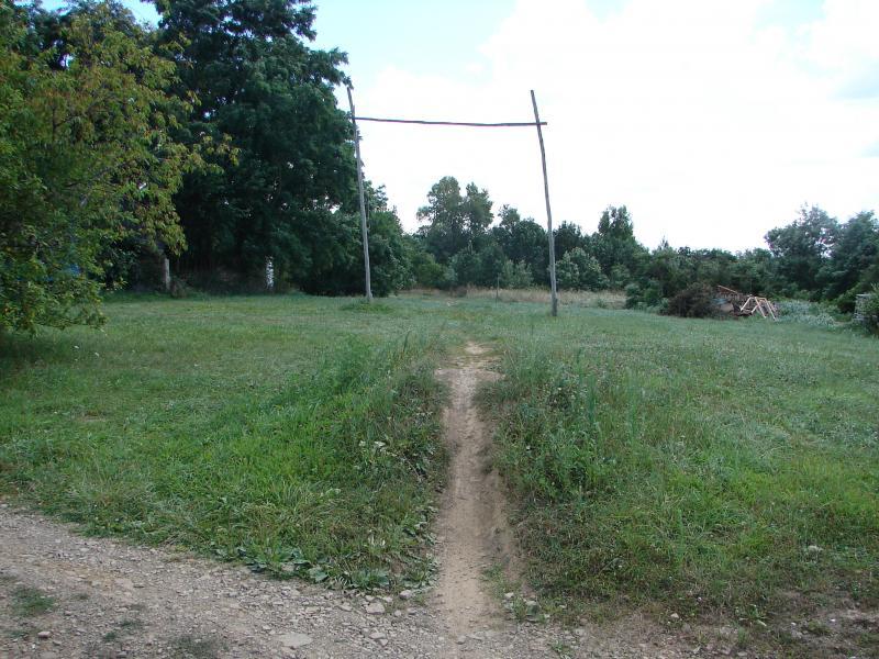 A grassy field with a dirt path leading through it, flanked by trees on either side. In the center, there is a wooden structure resembling a frame for a swing, with a horizontal beam supported by two vertical posts. In the background, more trees can be seen, and there is a pile of branches to the right. The sky is partly cloudy. Vultures Knob mountain bike trail.