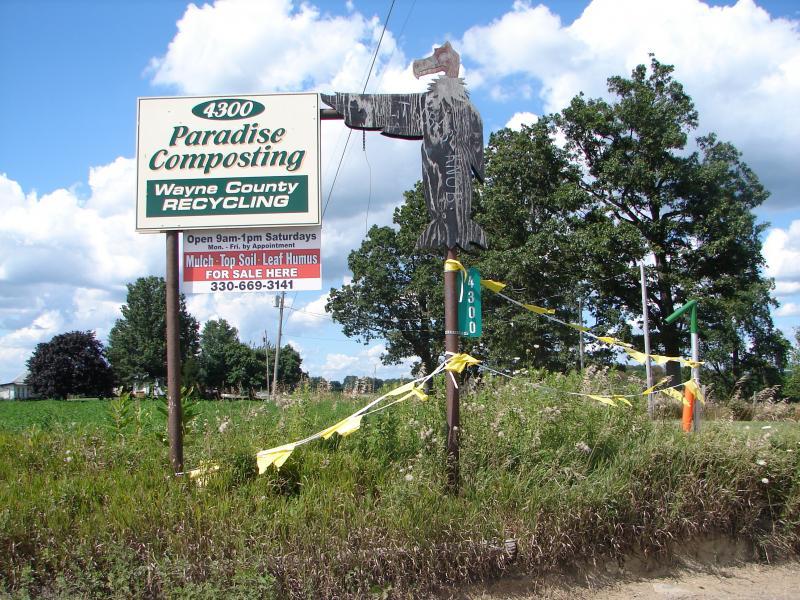 Sign for Paradise Composting and Wayne County Recycling, featuring a wooden sign with the business name and hours of operation, located at 4300. There are yellow caution ribbons around the area with additional information about mulch, top soil, and leaf humus for sale. The backdrop includes trees and a blue sky with clouds. Vultures Knob mountain bike trail.