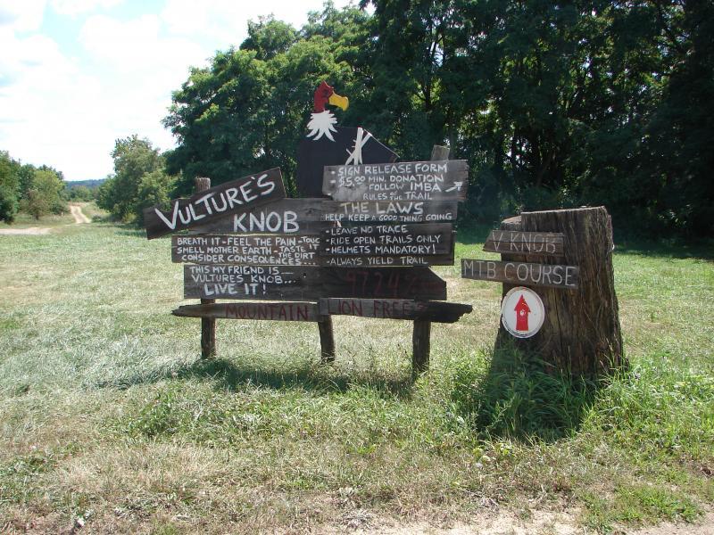 A wooden sign for Vulture's Knob, a mountain biking course, featuring various instructions and rules. The sign includes a rooster illustration at the top and text promoting safety, such as wearing helmets and following trail guidelines. The landscape in the background shows a dirt path and green trees, indicating a natural outdoor setting. Vultures Knob mountain bike trail.