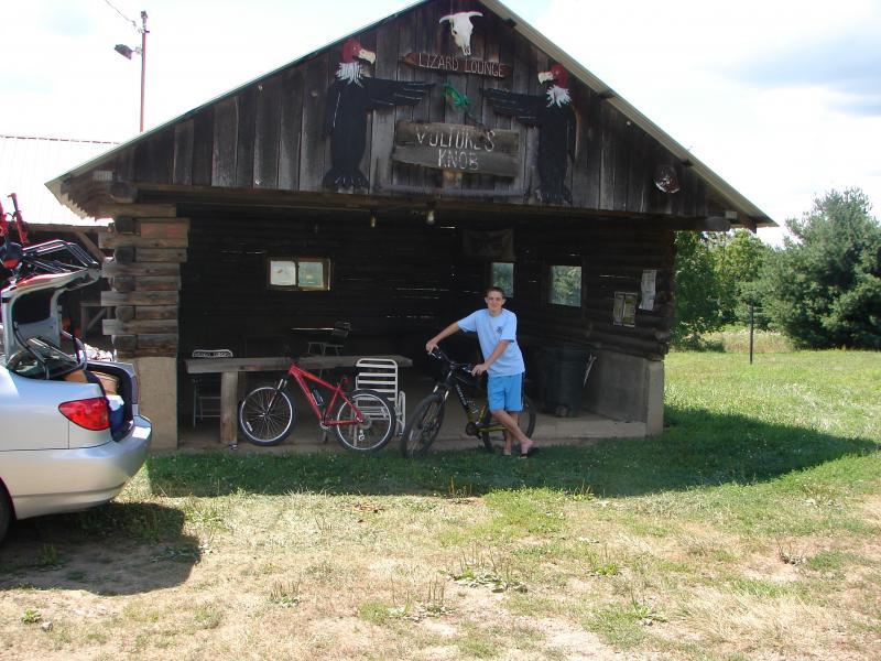 A young person stands next to a bicycle in front of a rustic wooden building labeled "Lizard Lounge" and "Vultures Knob." A silver car is parked nearby with bicycles visible on its roof. The scene is set in a grassy area with trees in the background under a blue sky. Vultures Knob mountain bike trail.