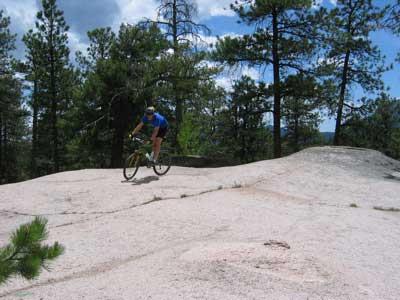 A mountain biker in a blue jersey rides across a smooth, rocky surface surrounded by pine trees under a partly cloudy sky. Buffalo Creek mountain bike trail.