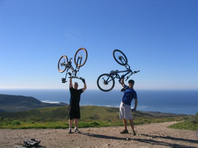 Two men are standing on a hillside with a scenic ocean view in the background, each holding their mountain bikes above their heads in celebration. The sky is clear and blue, and the landscape is lush with green hills. One man is wearing a black shirt and shorts, while the other is in a blue shirt and white shorts. Montana De Oro mountain bike trail.