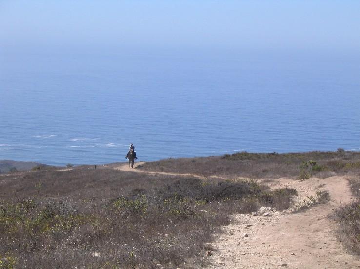 A person walking along a dirt hiking trail surrounded by grassy hills, with a panoramic view of the ocean and clear blue skies in the background. Montana De Oro mountain bike trail.