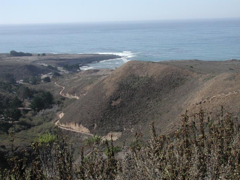 A scenic view of rolling hills overlooking the ocean, with waves gently breaking on the shore. A winding road can be seen in the foreground, leading through the landscape. The horizon is clear, and the sky is bright, suggesting a sunny day. Montana De Oro mountain bike trail.