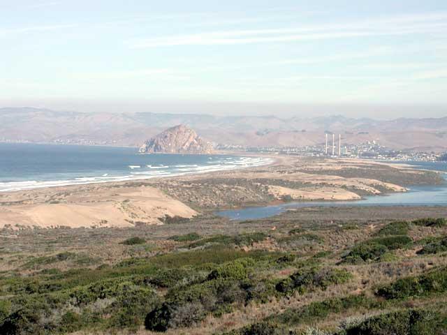 A scenic view of a coastal landscape featuring a sandy beach, calm ocean waves, and distant mountains. In the foreground, lush greenery is visible, leading down to a winding river. A prominent rock formation stands out in the ocean, with a small town and tall structures visible along the shoreline in the background. The sky is clear with soft clouds, suggesting a tranquil atmosphere. Montana De Oro mountain bike trail.